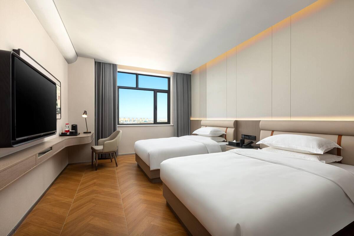 A hotel room with two beds, a TV, a desk with a lamp, and a view of the city.