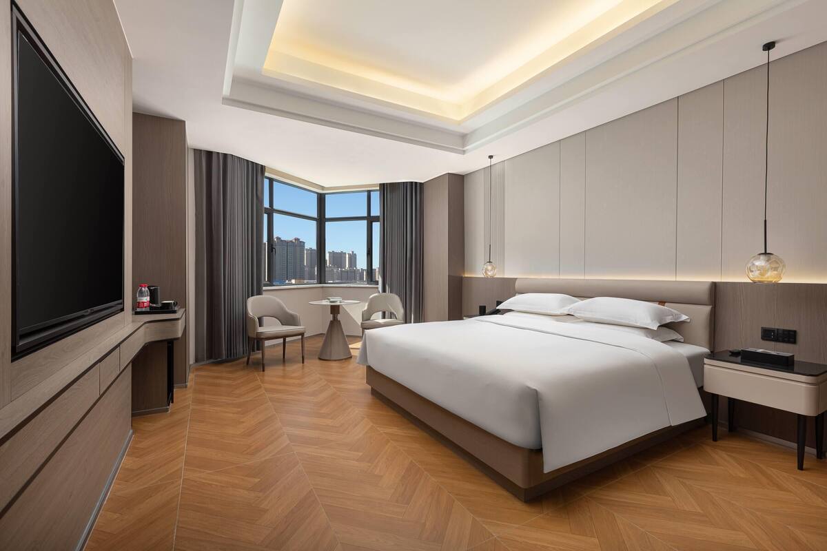 A modern hotel room with a large bed, a flat-screen TV, a desk, and a view of the city.