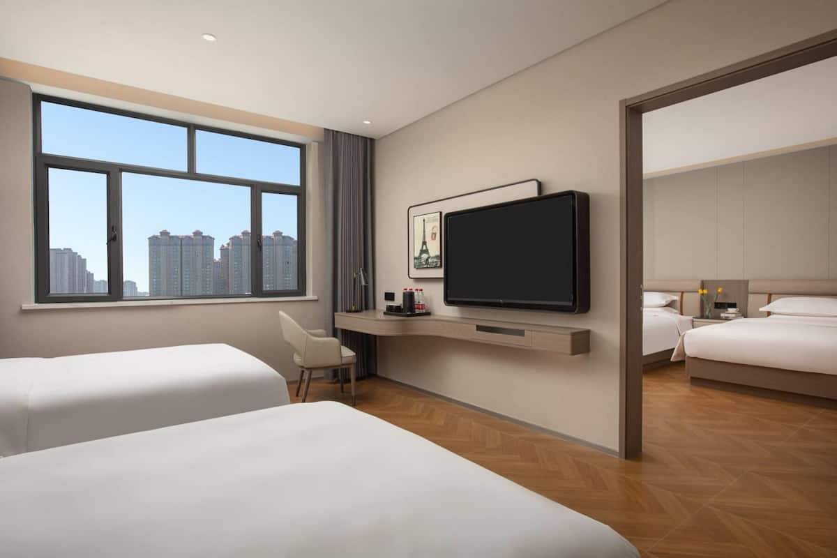 A hotel room with two beds, a desk, a TV, and a large window with a city view.
