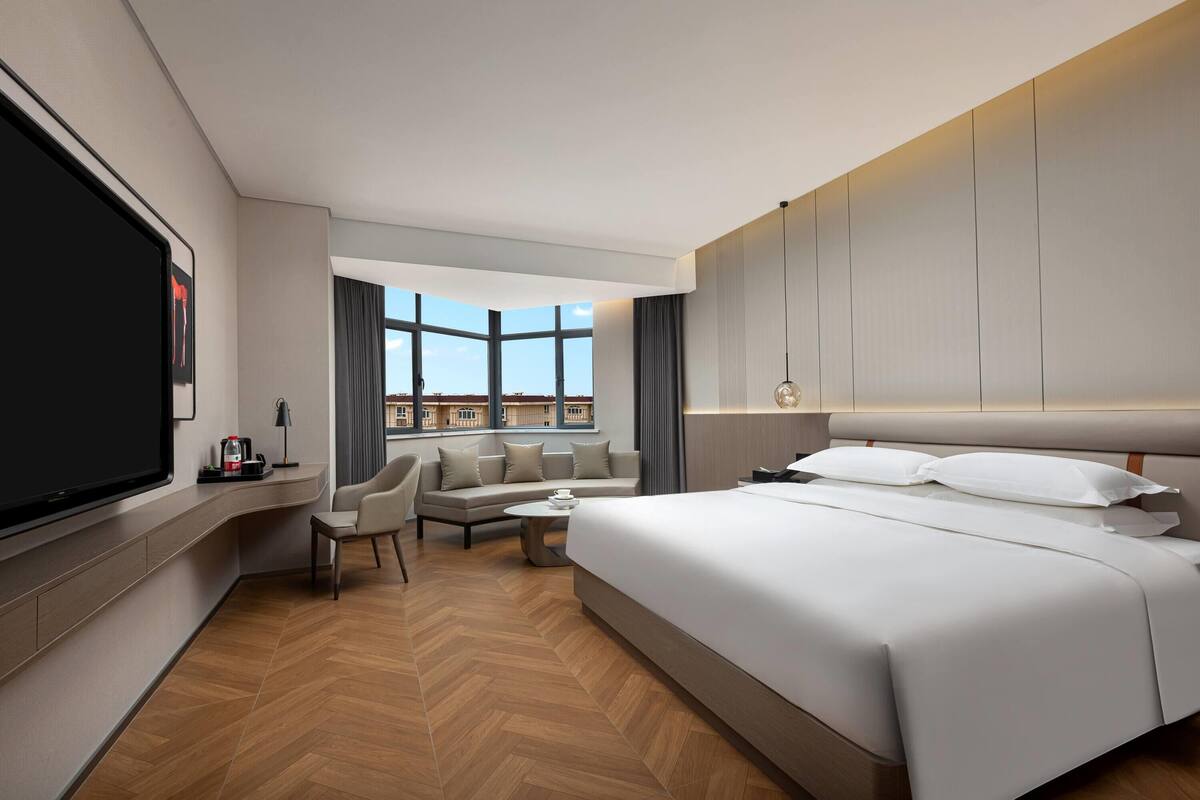 A modern hotel room with a large bed, a sofa, a TV, and a view of buildings through large windows.