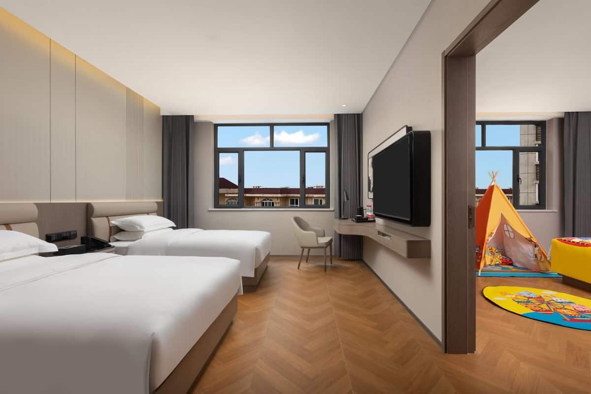 A hotel room with two beds, a TV, a window with a view, and a play area with a tent and toys.