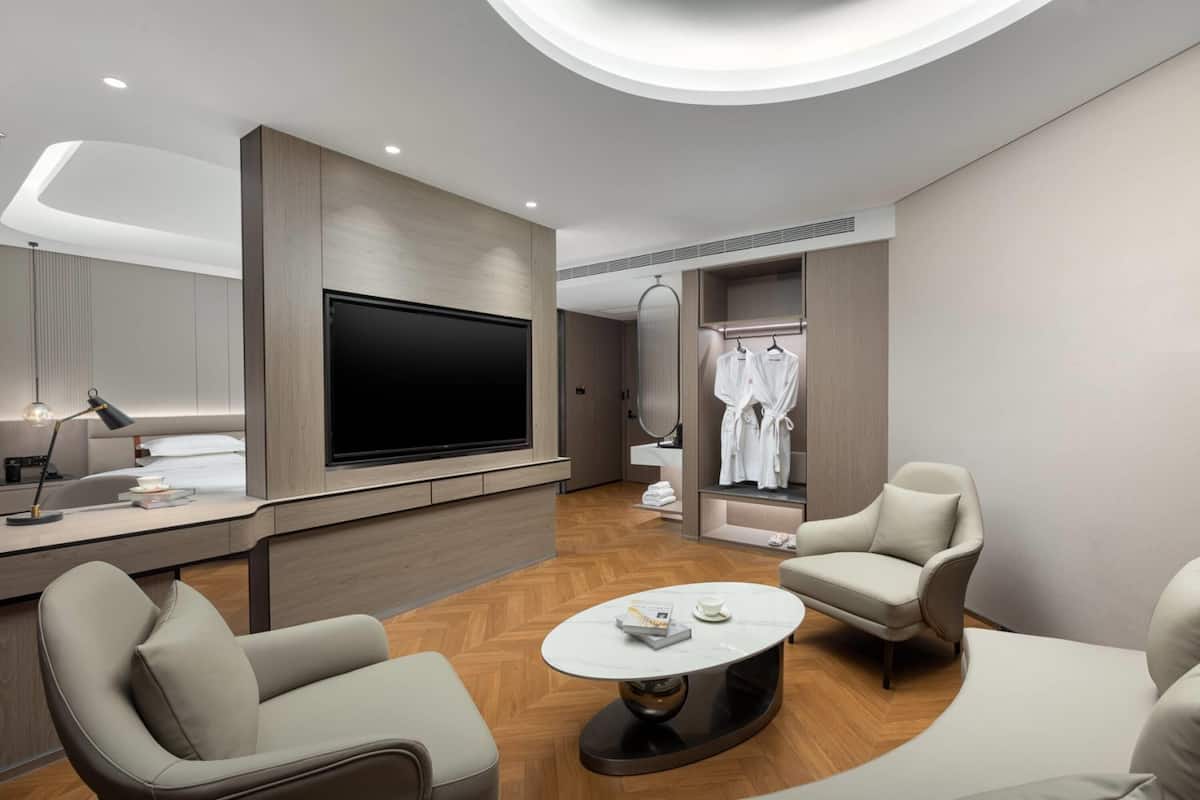 A modern hotel room with a flat-screen TV, a desk with a lamp, a round coffee table, two armchairs, a walk-in closet with hanging robes, and a wooden floor.