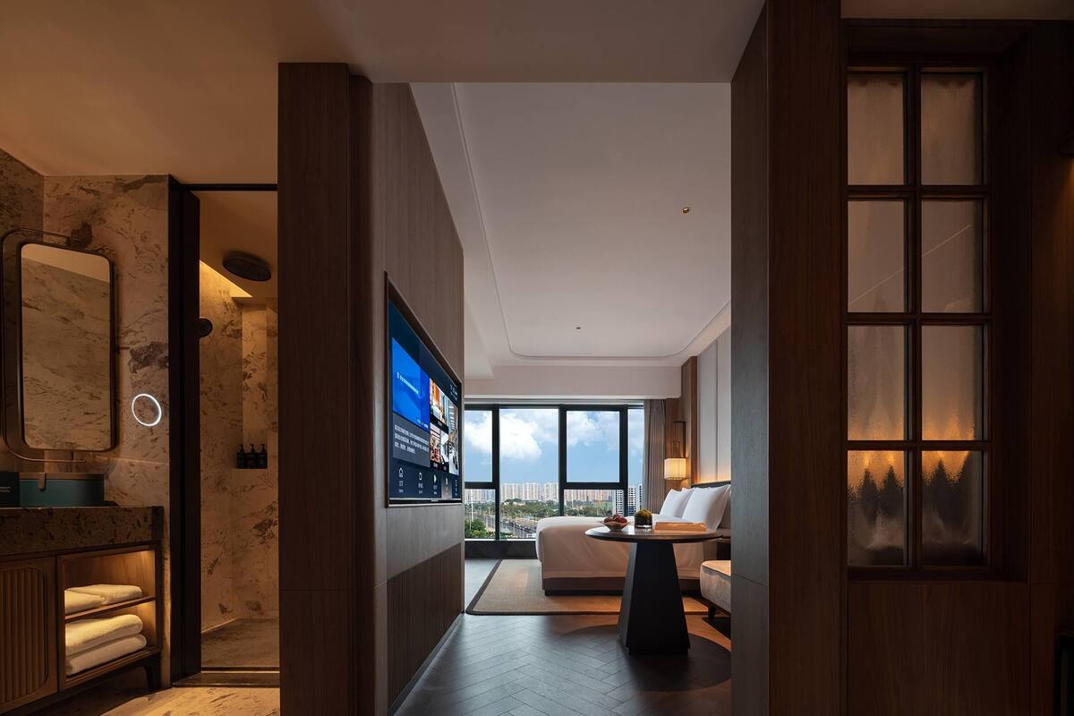 A modern hotel room with a large window, a flat-screen TV, a round table, and a cozy seating area.