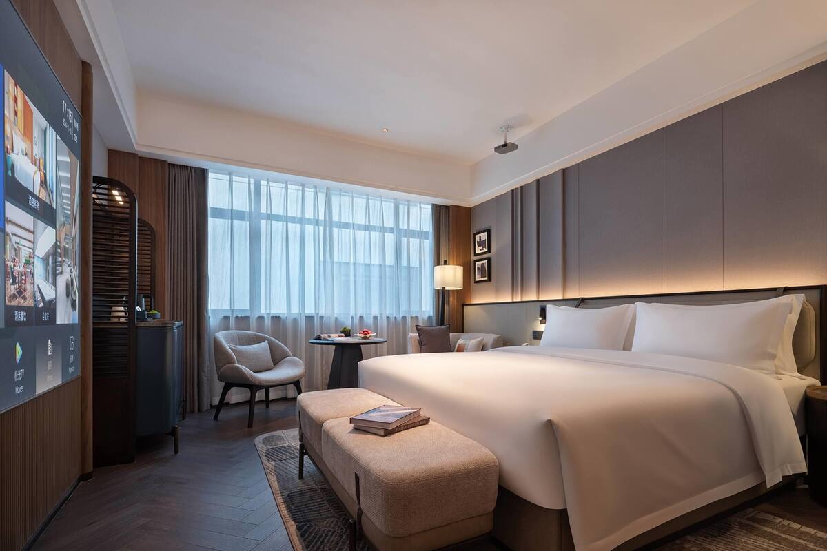 A hotel room with a large bed, a desk, a chair, a small table with a lamp, and a large window with curtains.
