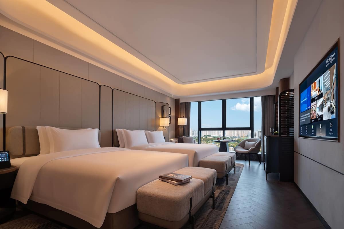 A modern hotel room with two beds, a TV on the wall, and a city view.