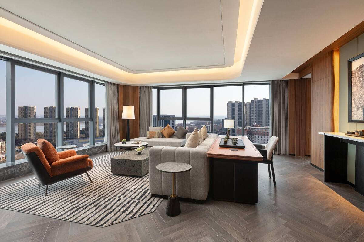 A modern hotel room with a large window, a sofa, a dining table, and a small round table.