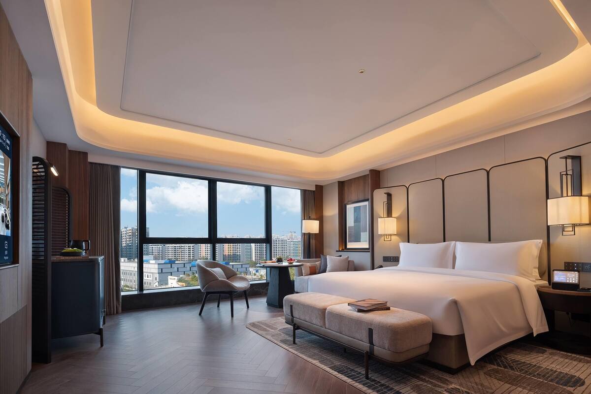 A modern hotel room with a large bed, a desk, a chair, and a view of the city.