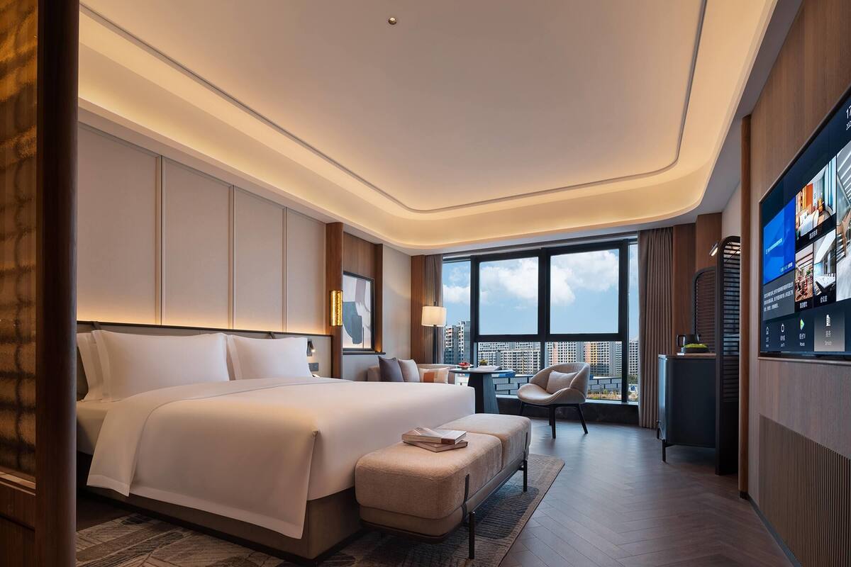 A modern hotel room with a large bed, a seating area, a TV, and a city view.