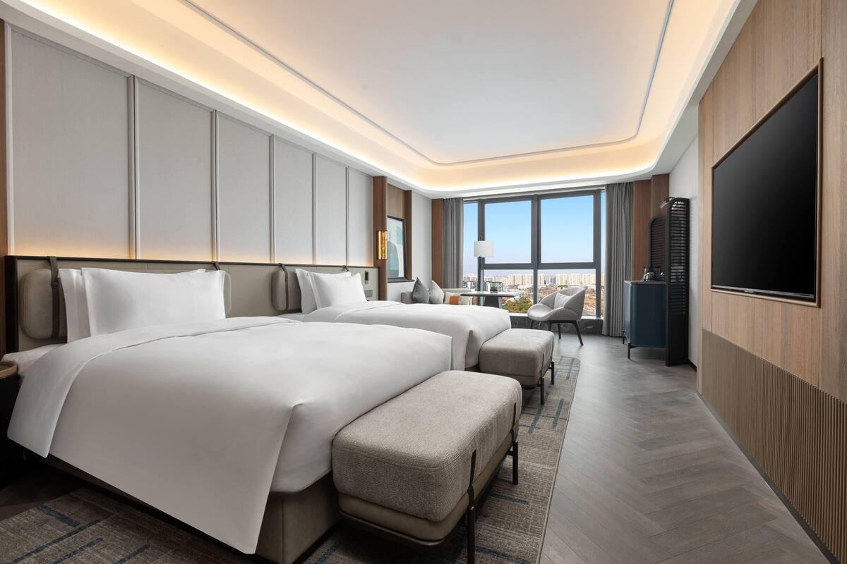 A modern hotel room with two large beds, a flat-screen TV, and a city view.