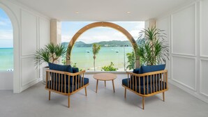 A room with a large arched window overlooking a beach, two wooden chairs with blue cushions, and a small round table.