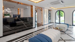 A modern hotel lobby with a marble floor, a reception desk, seating area, and large windows offering a view of greenery.