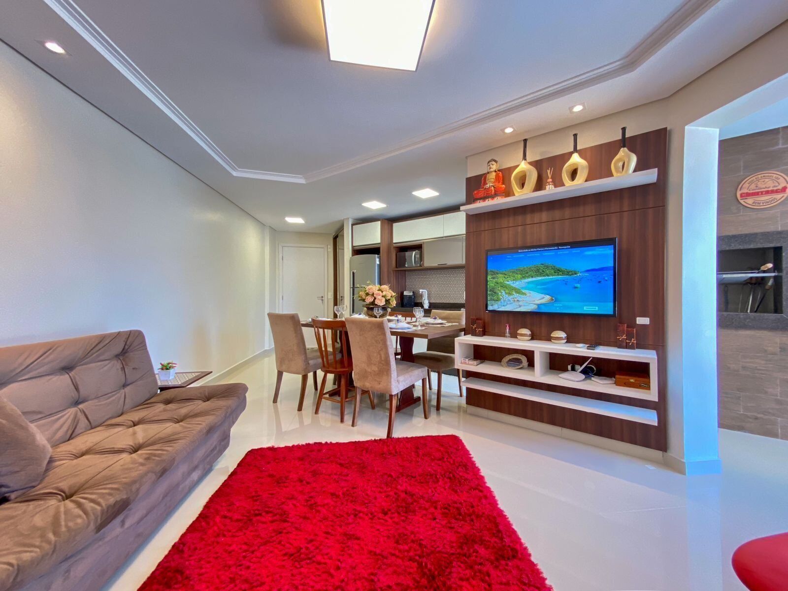 Una sala de estar moderna con sofá, mesa de comedor, sillas, alfombra roja, televisor montado en una pared de madera y una pequeña cocina.