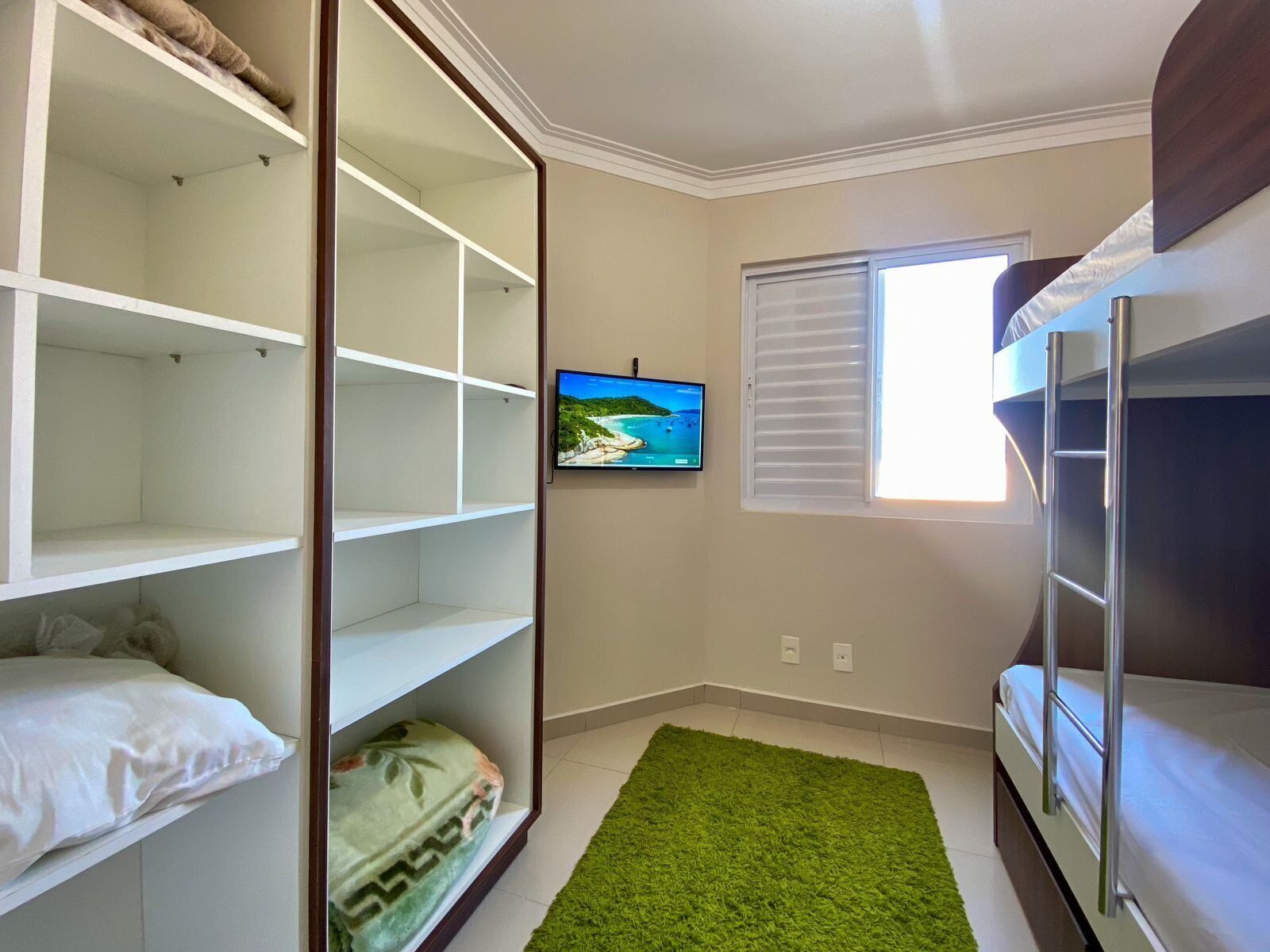 Um quarto com uma cama beliche, um tapete verde, uma televisão na parede e uma janela com persianas.