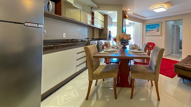 A modern kitchen with a stainless steel refrigerator, a dining area with a wooden table and chairs, and a red rug. - Resid. Clube Mar Grosso-Laguna - LAG605 (Laguna)