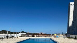 A rectangular swimming pool with a tiled deck, surrounded by a fence and a building. - Resid. Clube Mar Grosso-Laguna - LAG605 (Laguna)
