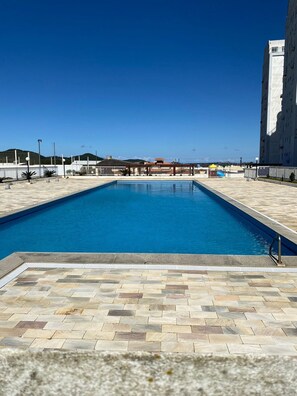 A rectangular swimming pool with a tiled deck, surrounded by a fence and a building. - Resid. Clube Mar Grosso-Laguna - LAG605 (Laguna)