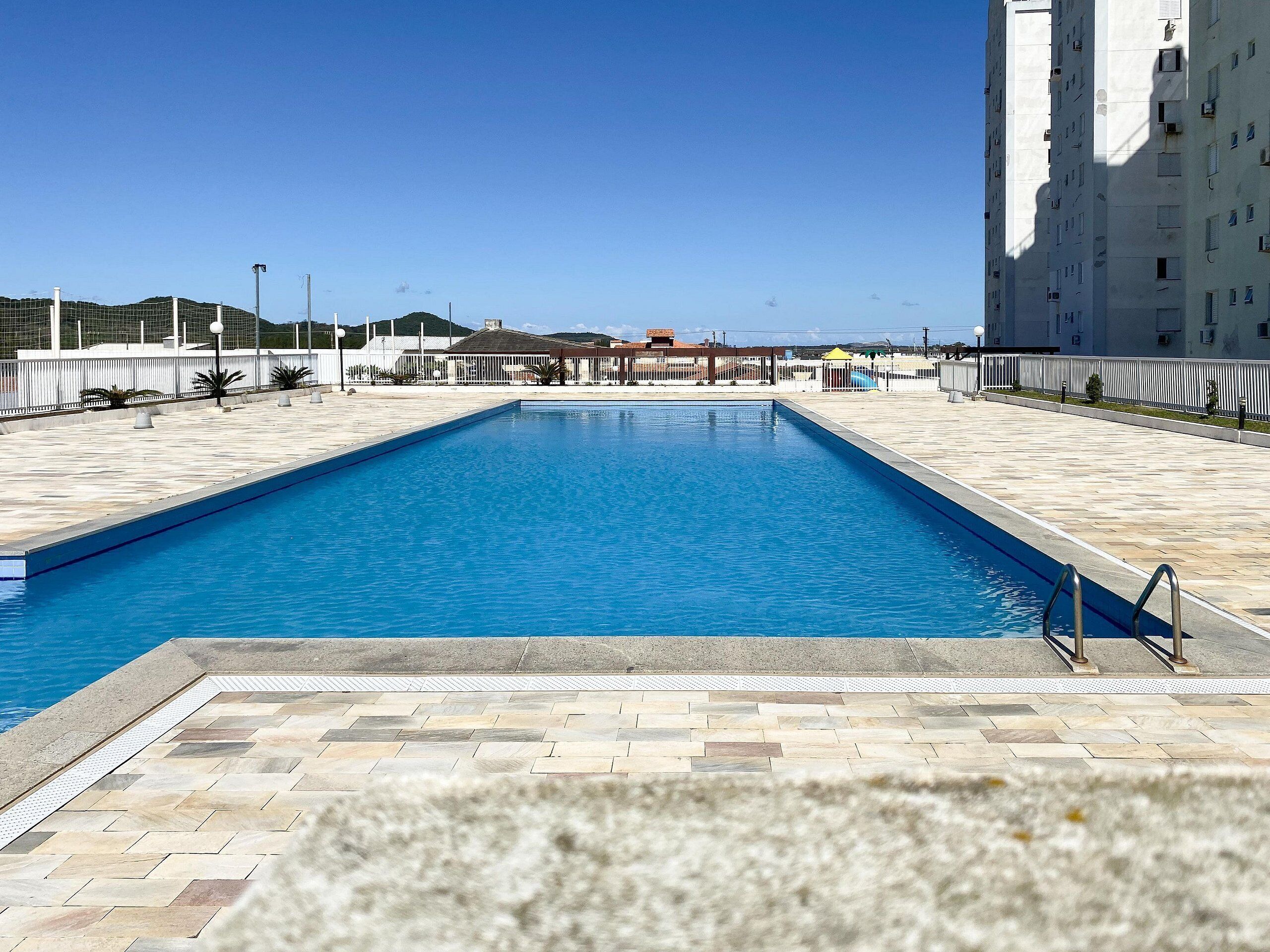 Una piscina rectangular con un borde empedrado, una escalera metálica y un cielo azul despejado al fondo.