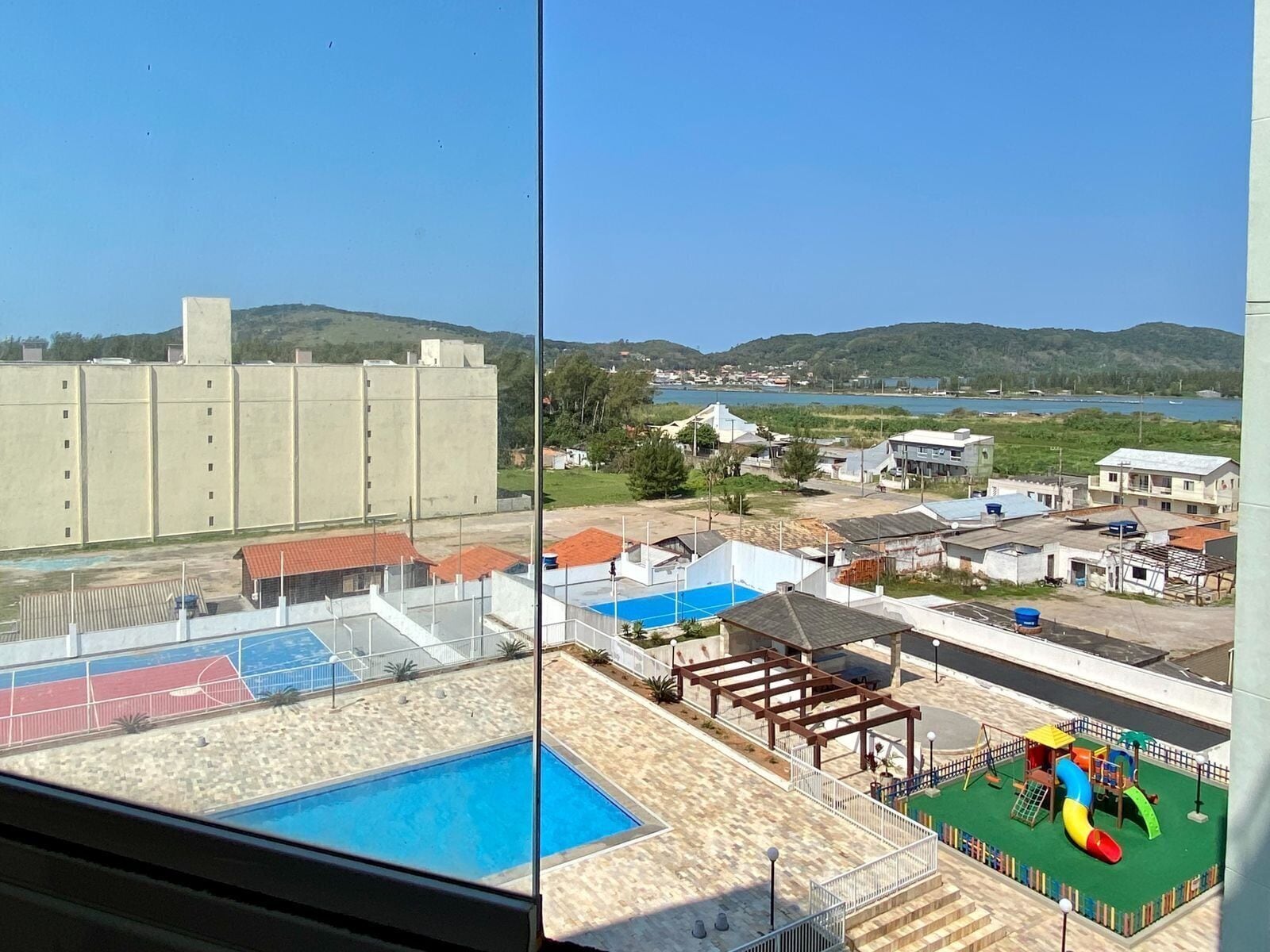 Vista de uma piscina, área de recreação infantil e quadra esportiva vista da janela.