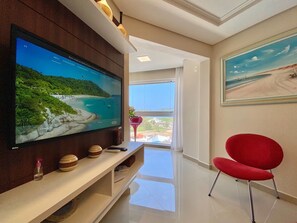 A modern living room with a large flat-screen TV on a wooden entertainment unit, a red chair, and a painting of a beach scene on the wall. - Resid. Clube Mar Grosso-Laguna - LAG605 (Laguna)