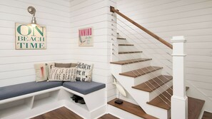 Una escalera con escalones de madera y barandilla blanca, un banco con cojines y adornos en la pared, incluyendo un cartel que dice 'ON BEACH TIME'.