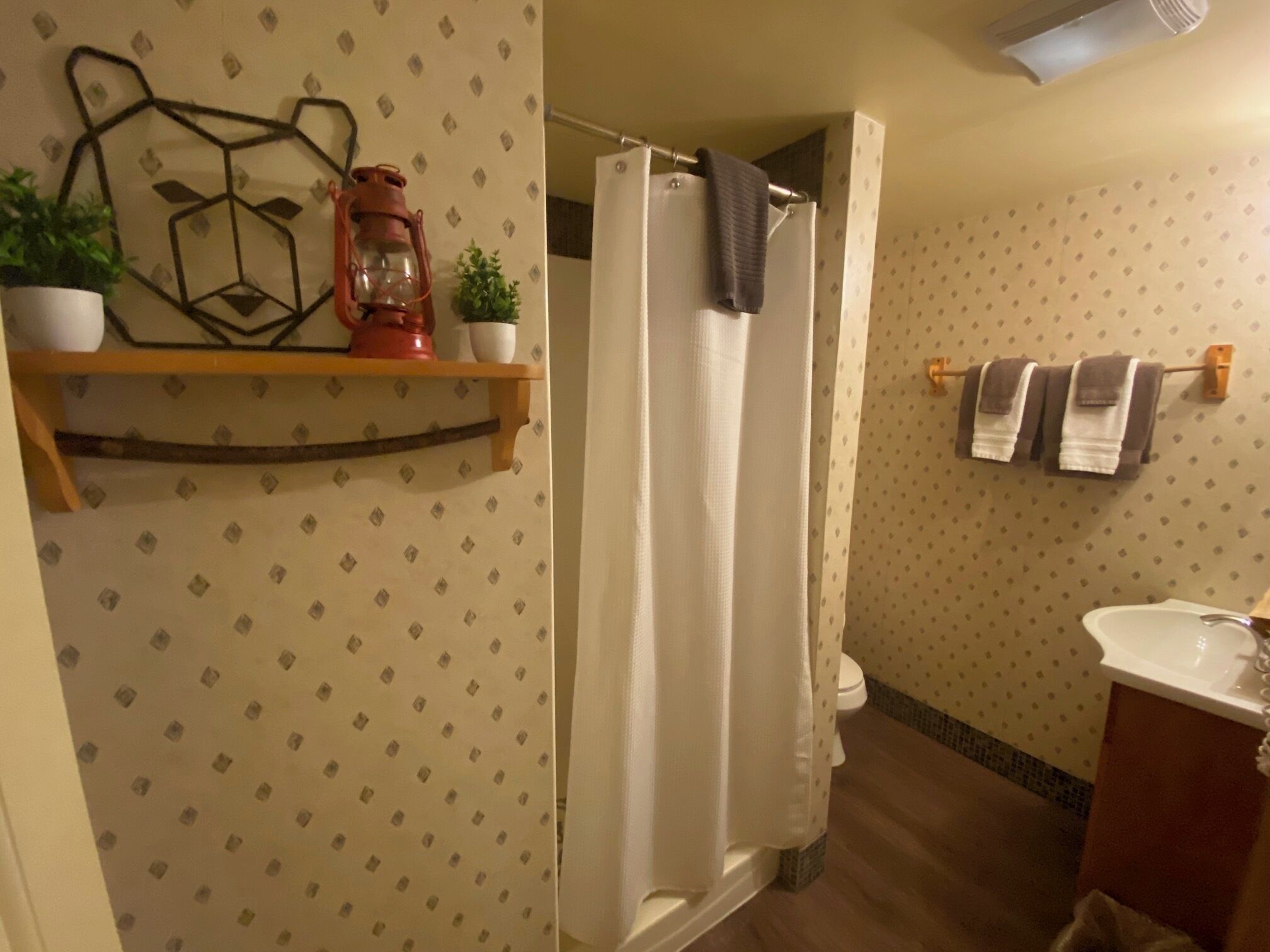 A bathroom with a shower curtain, a toilet, a sink, and a shelf with plants and a lantern.