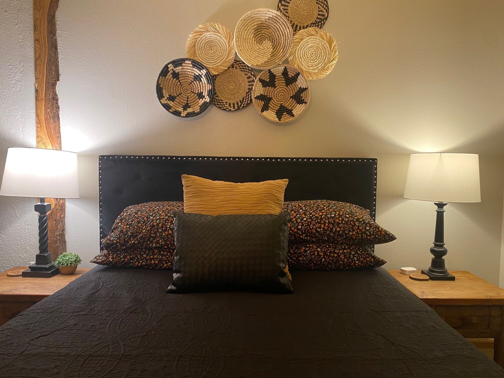 Un lit recouvert d’une couvre-lit sombre, deux lampes et un mur décoré de paniers tressés.