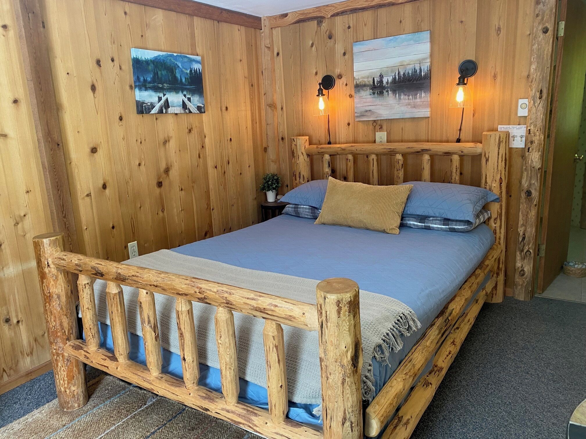 Une pièce aménagée comme un chalet en bois, avec un lit, deux lampes fixées au mur et deux tableaux.