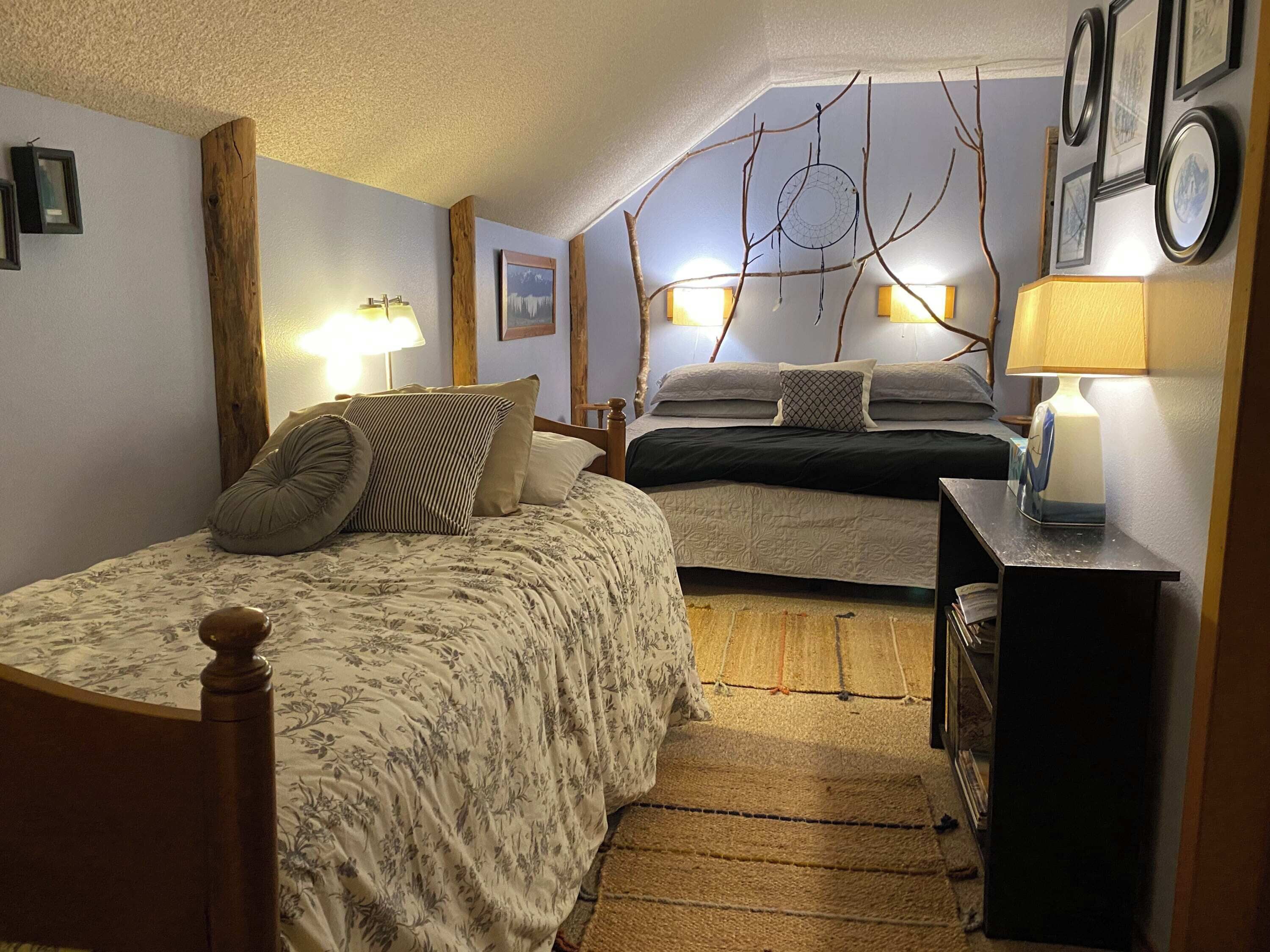 A bedroom with two beds, wooden headboards, a small bedside table with a lamp, and a wall-mounted dreamcatcher.