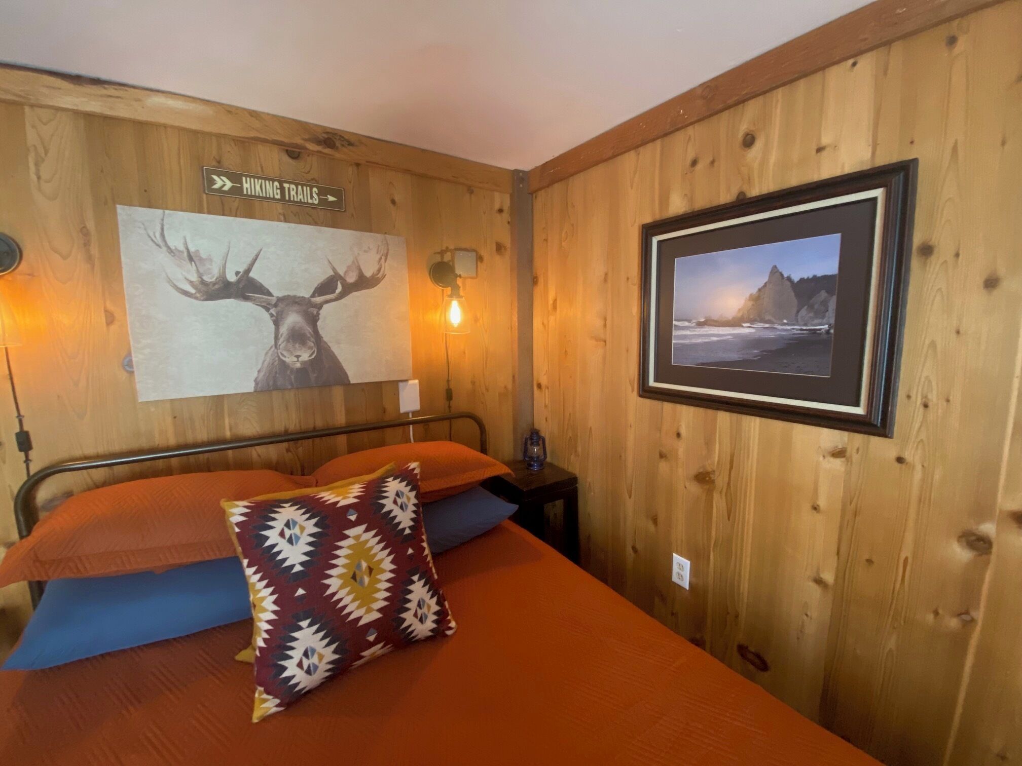 A wooden room with a bed, a sign indicating hiking trails, a wall-mounted light, and a framed picture of a coastal scene.