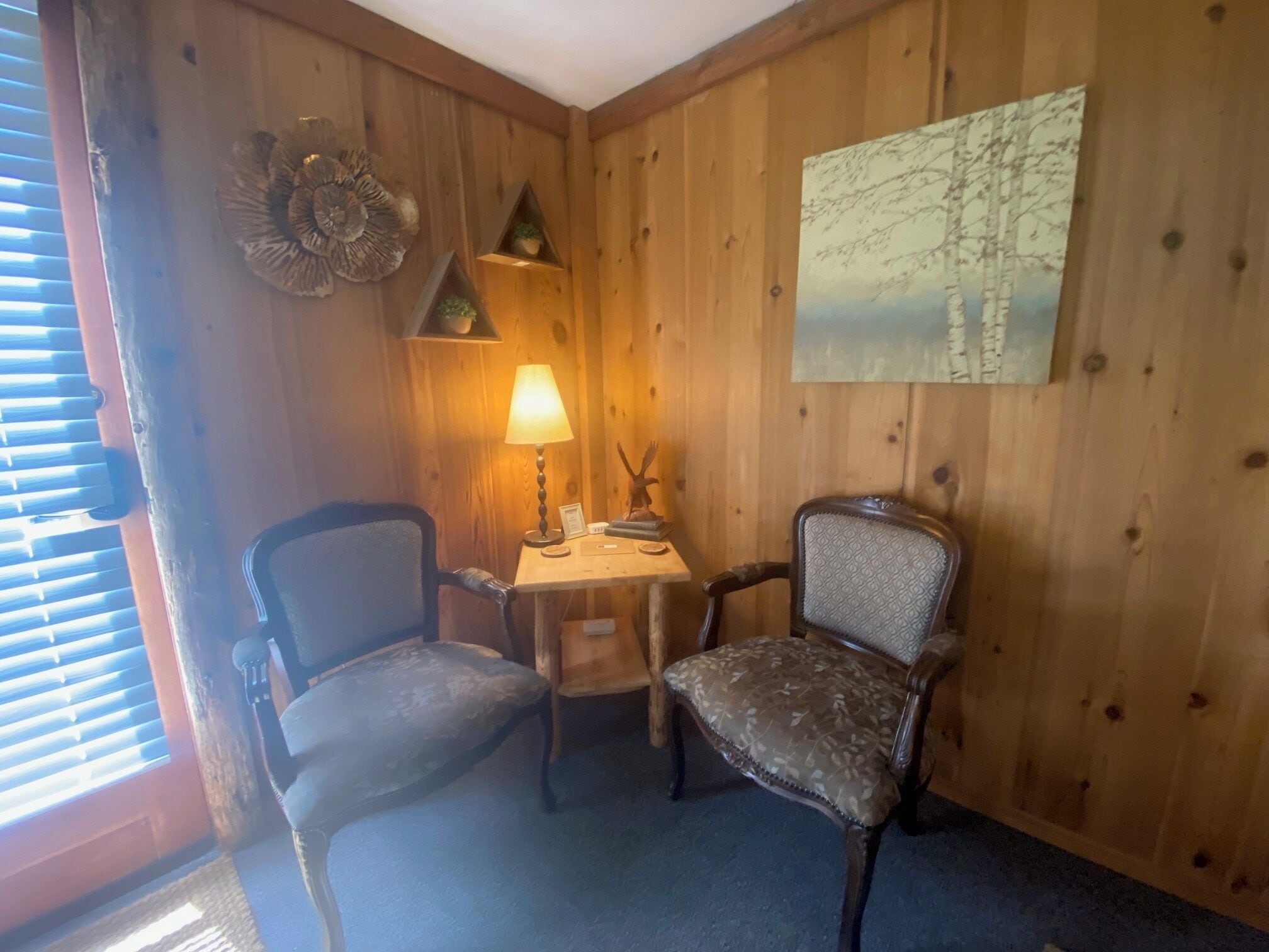 A wooden-paneled room with two chairs, a small table with a lamp, and a wall-mounted shelf with decorative items.