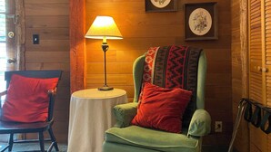 A cozy room with wooden walls, a green armchair with a red pillow, a small round table with a lamp, and two framed floral pictures on the wall. - Manitou Lodge (Forks)