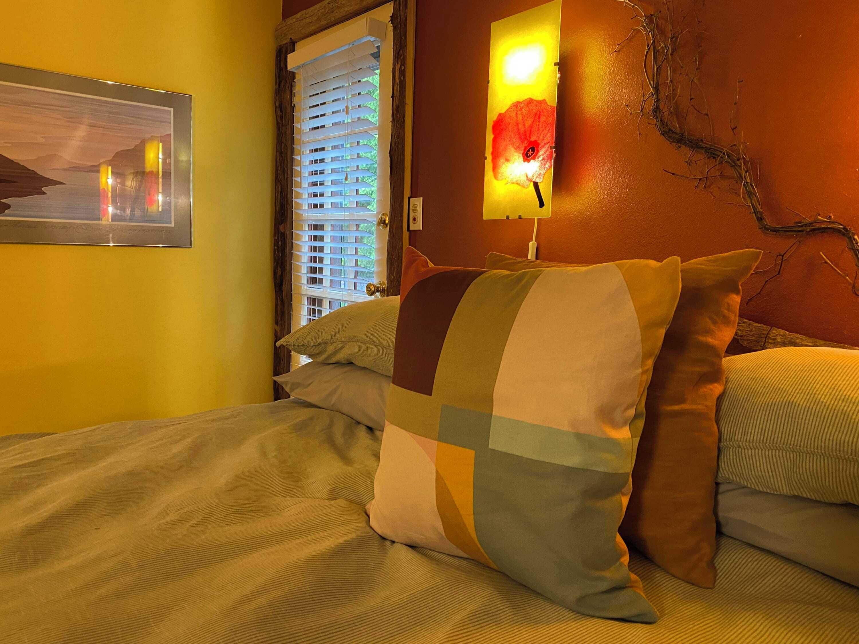 A bed with multiple pillows, a painting on the wall, and a window with blinds.