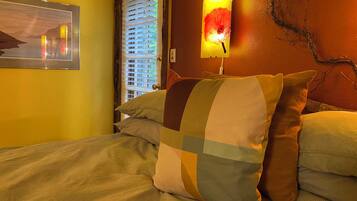 A bed with multiple pillows, a painting on the wall, and a window with blinds.