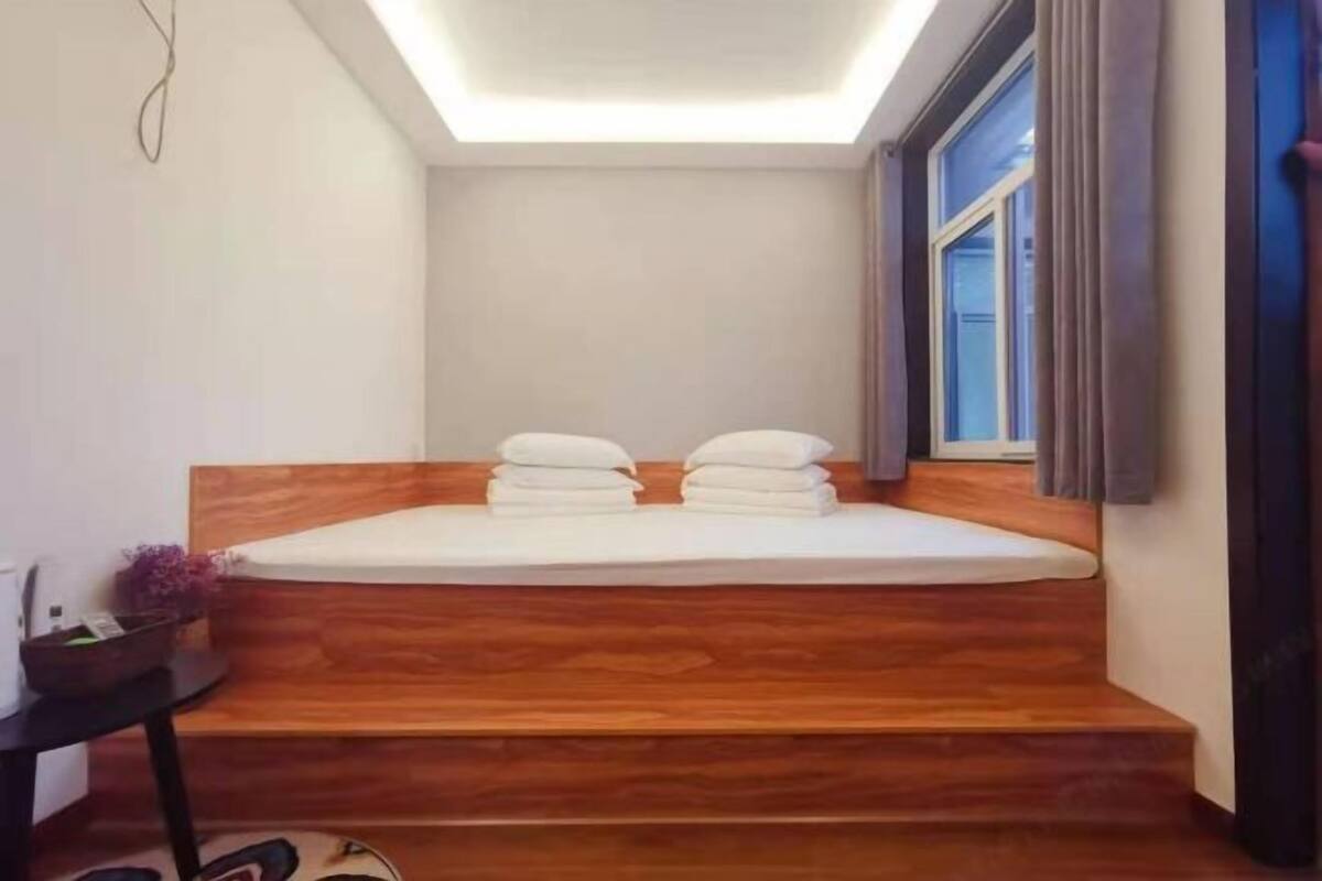 A room with a wooden headboard, a bed with white linens, a small table with a plant, and a window with curtains.
