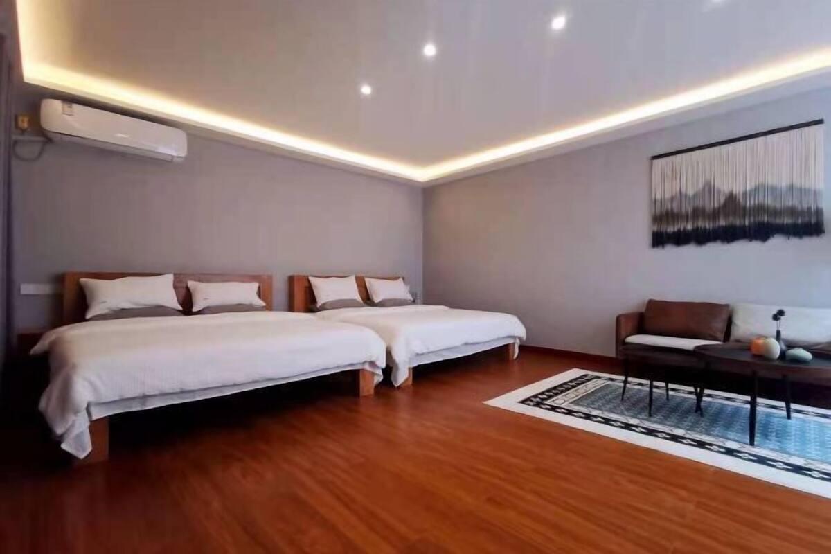 A hotel room with two beds, a small table, a sofa, and a wall-mounted air conditioner.