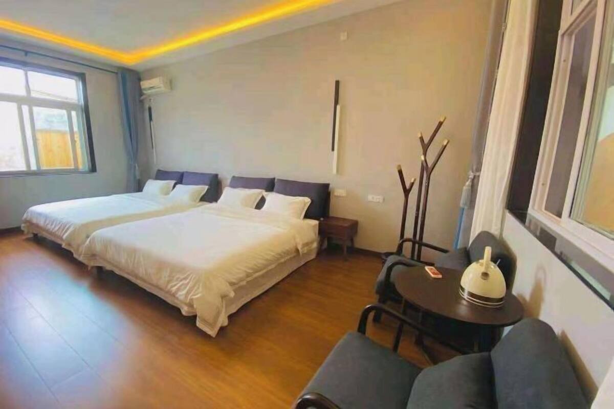 A hotel room with two beds, a small table with a kettle, a chair, and a window.