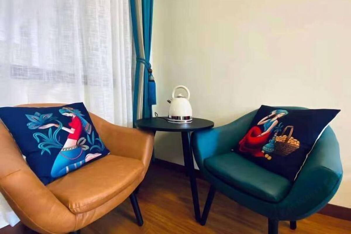 Two chairs with distinct pillows, a white kettle on a small table, and a window with curtains.