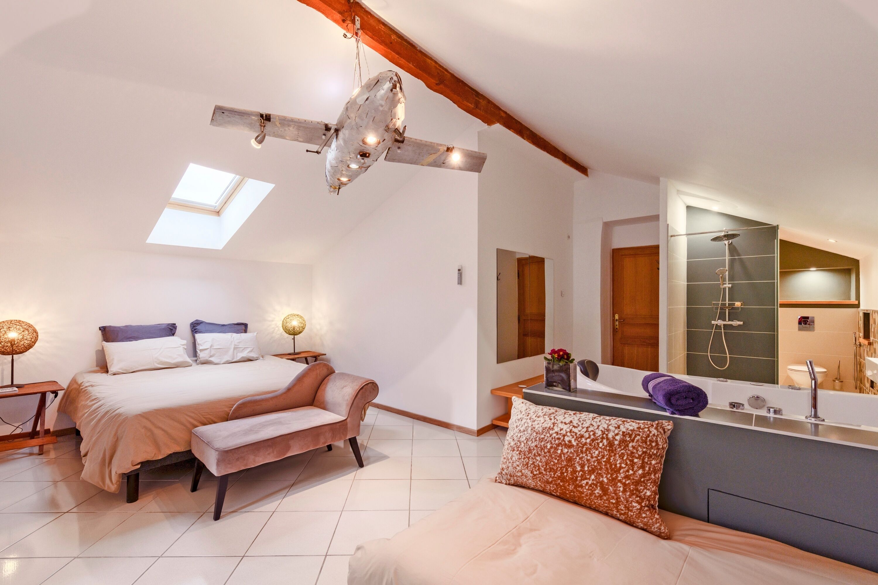 A bedroom with a bed, bench, skylight, and a bathroom with a shower and mirror.
