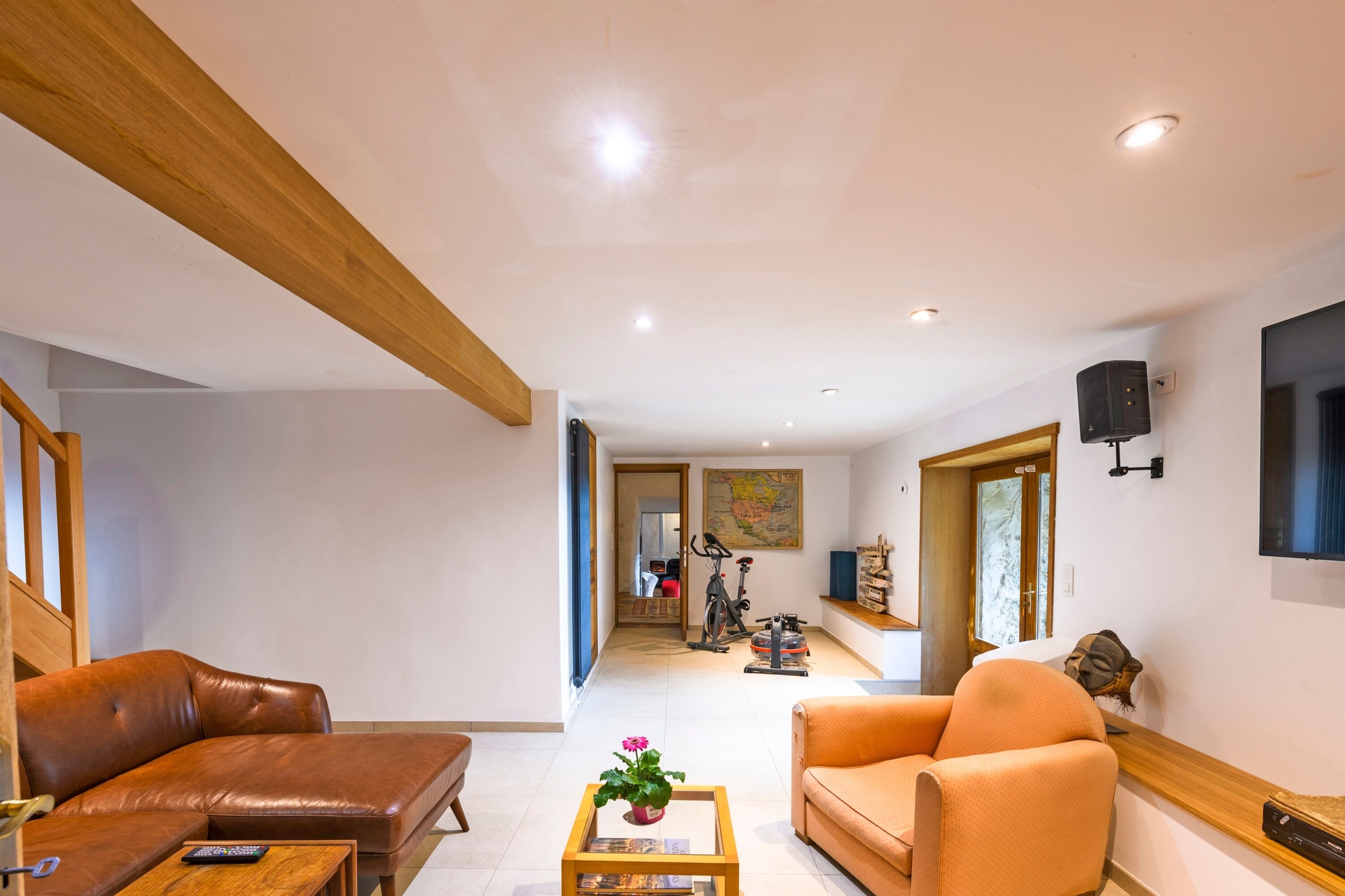 A spacious living area with a brown leather sofa, an orange armchair, and a wooden coffee table with a flower vase. There is a television mounted on the wall and a bicycle in the background.