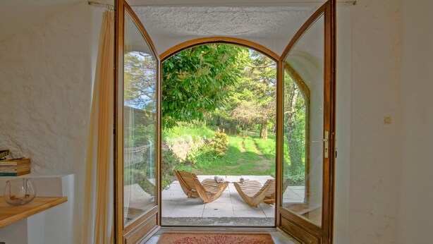 Ein Zimmer mit Glastür und Zugang zu einem Außenbereich mit zwei Holzstühlen und Blick auf einen Garten.