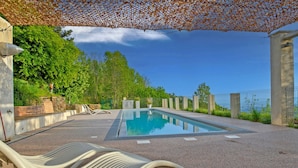 Ein Schwimmbecken mit glasklarer, blauer Wasserfläche, umgeben von einem Kiesgrund und einer Pergola mit orangefarbenen Blättern.