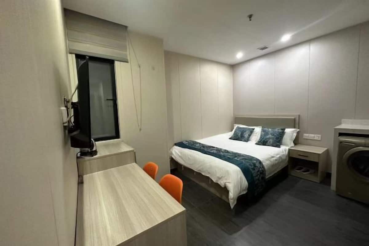 A modern hotel room with a bed, a desk, a chair, a washing machine, and a mirror.