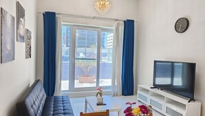 A modern living room with a sofa, a dining table with a vase of flowers, a TV on a stand, and a view of the city through large windows.
