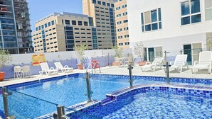 A rooftop pool with blue mosaic tiles, surrounded by white lounge chairs and potted plants.