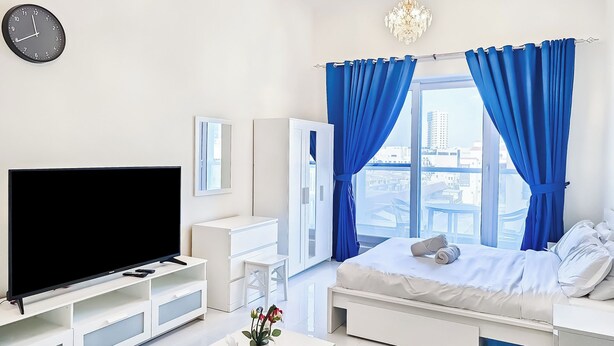 A modern bedroom with a TV, white furniture, a bed with blue curtains, and a city view.