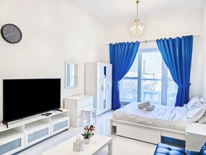 A modern bedroom with a TV, white furniture, a bed with blue curtains, and a city view. - MK Residence (Dubai)