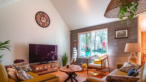 A cozy living room with a TV, wooden furniture, a round table, and a large window with a view of greenery. - Le Chêne Sacré - Piscine intérieur (Plouay)