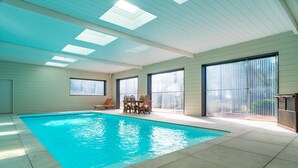 An indoor swimming pool with tiled flooring, a wooden table with chairs, and large windows with blinds. - Le Chêne Sacré - Piscine intérieur (Plouay)
