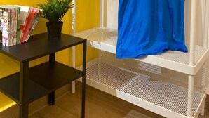 A room with a yellow wall, a white metal shelf, a blue sports jersey hanging on the shelf, and a small table with a plant and books. - Stay Costa House Naples (Naples)