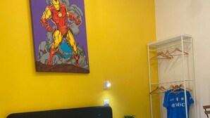 A bedroom with a bed, a yellow wall, a painting of a superhero, and a clothing rack with a blue shirt. - Stay Costa House Naples (Naples)
